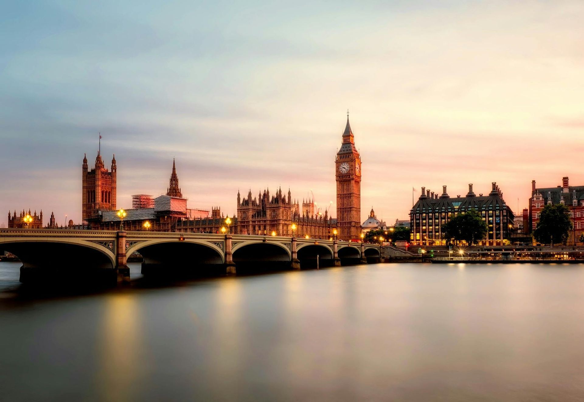 The Houses of Parliament and Big Ben illuminated against a soft, sunset sky over the River Thames in London.