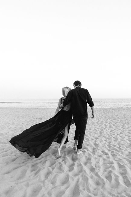 A couple in dark clothing walking hand-in-hand along a sandy beach towards the ocean, captured in black and white.
