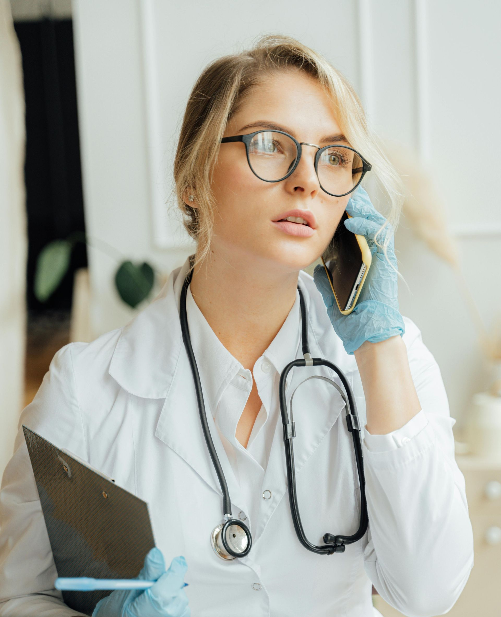 A healthcare worker wearing a white coat, stethoscope, and blue gloves holds a clipboard while speaking on a phone.