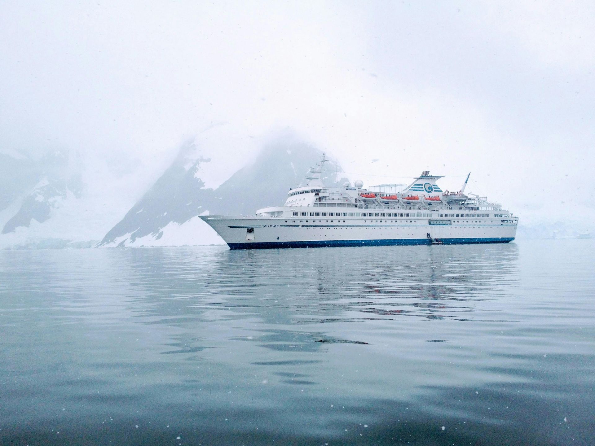 A white cruise ship floats in calm water surrounded by snowy, fog-covered mountains in an Antarctic landscape.