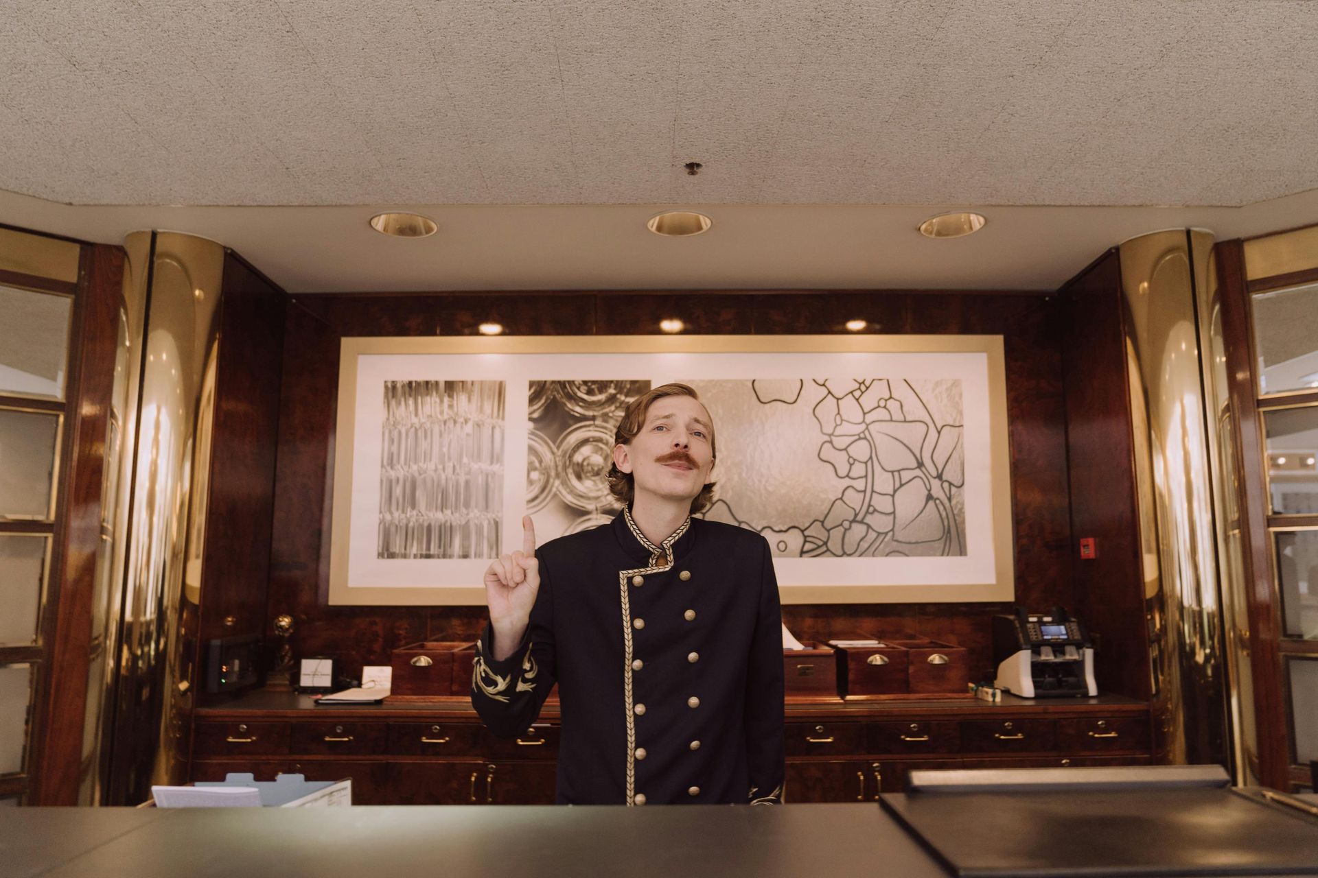 A hotel staff member in a dark uniform with gold trim stands behind a wooden desk, gesturing with one raised finger.