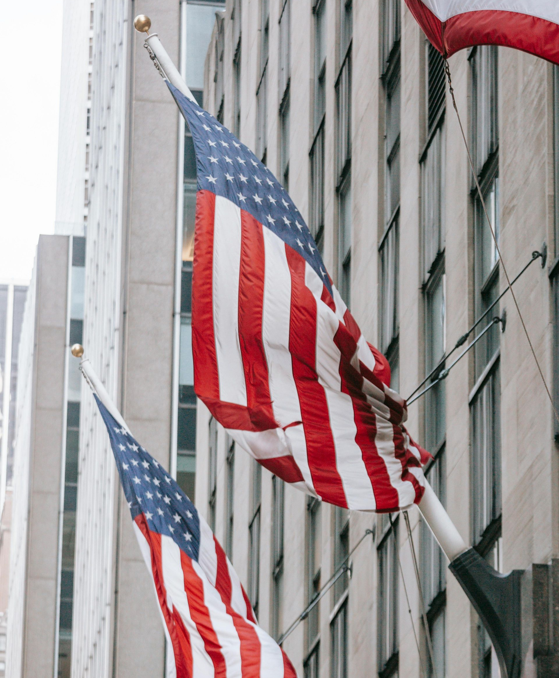 Two American flags fly against the side of a tall stone building.