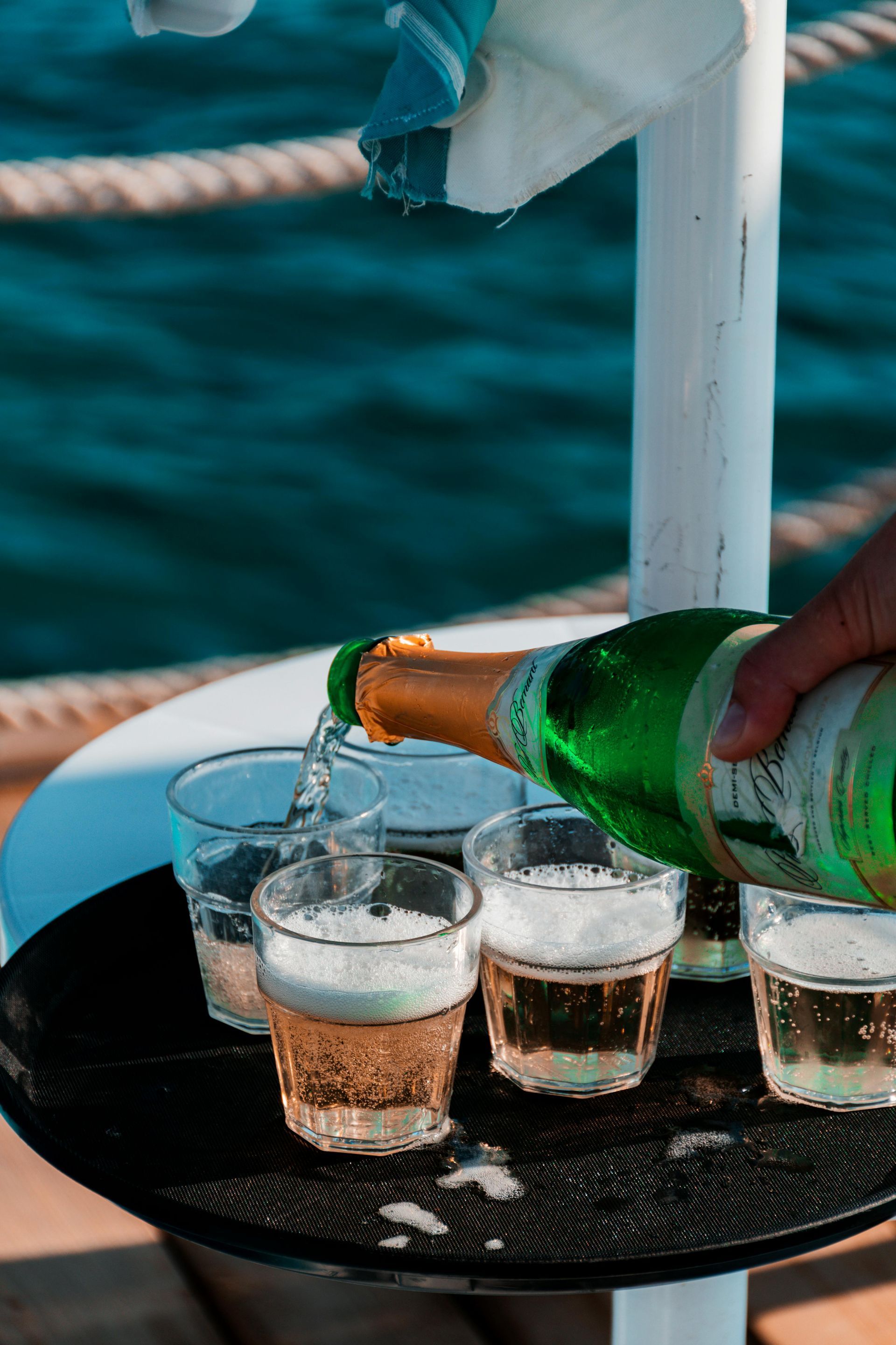 A hand pours sparkling wine from a green bottle into several glasses arranged on a black tray against a blue water backdrop.