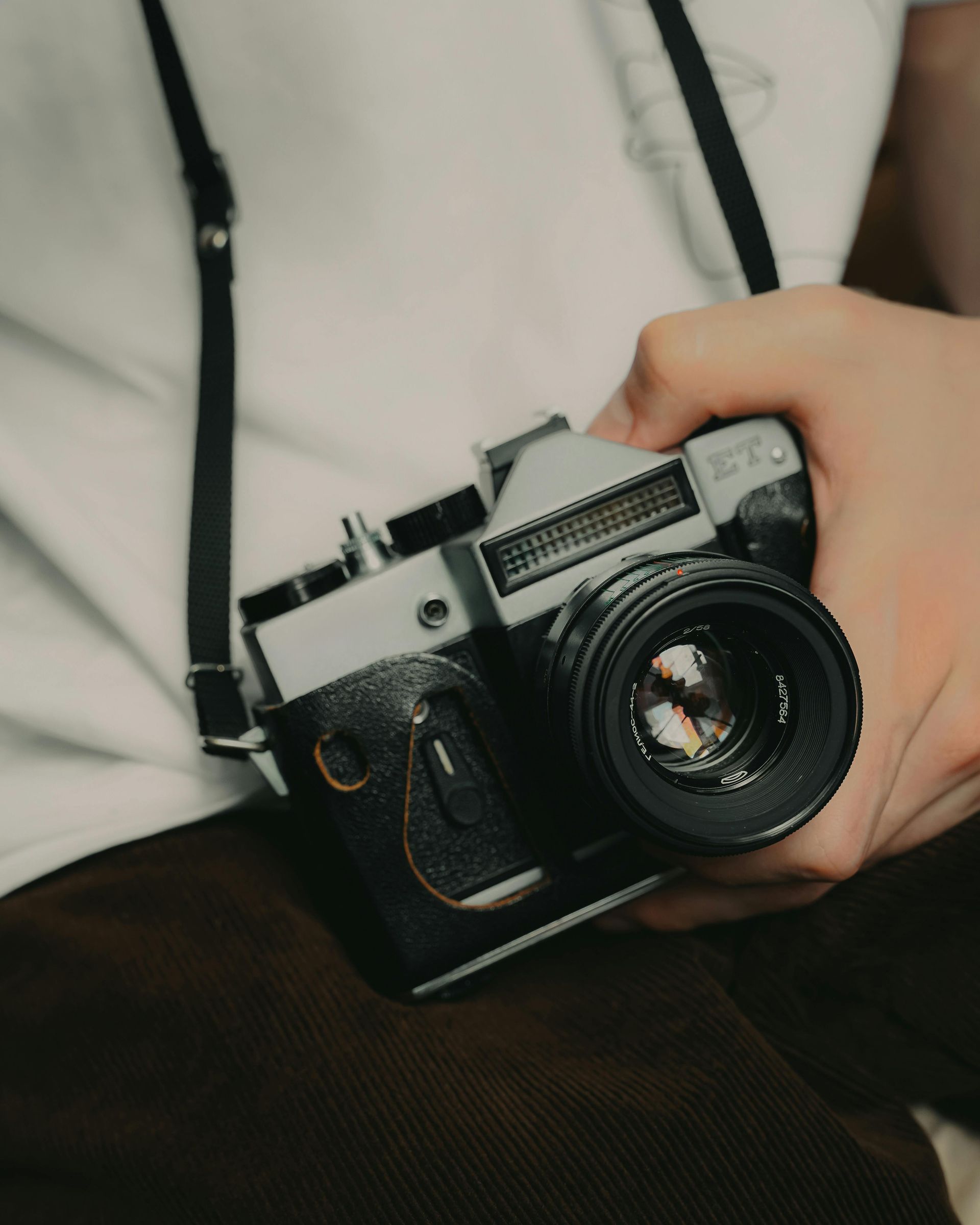 A close-up of hands holding a vintage silver and black SLR film camera with a strap, set against a light shirt and pants.