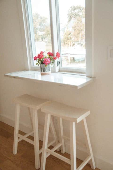 Two white stools are sitting under a window with a vase of flowers on it.