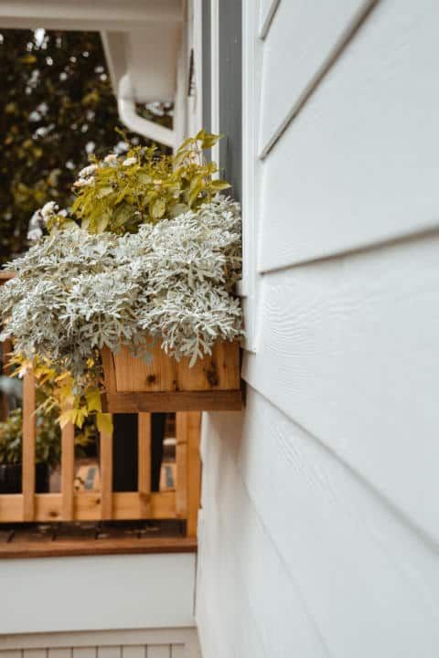 A wooden box filled with plants is hanging on the side of a house.
