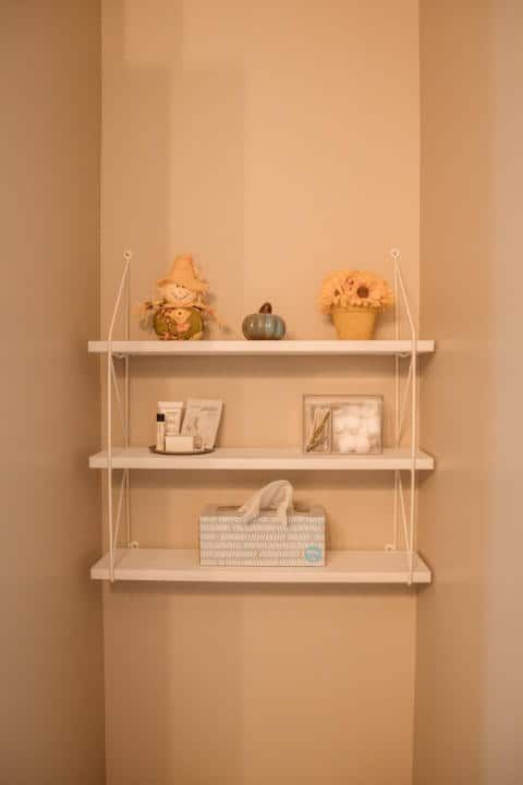 A white shelf in a corner of a room with flowers and pumpkins on it.