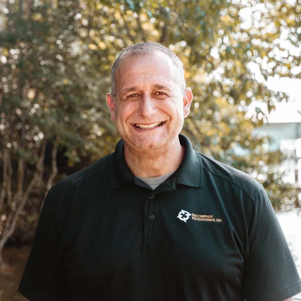 A man in a black shirt is smiling for the camera while standing in front of trees.