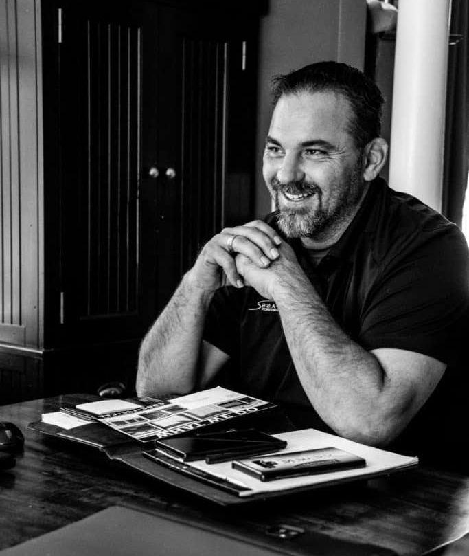 A man is sitting at a table with his hands folded and smiling.