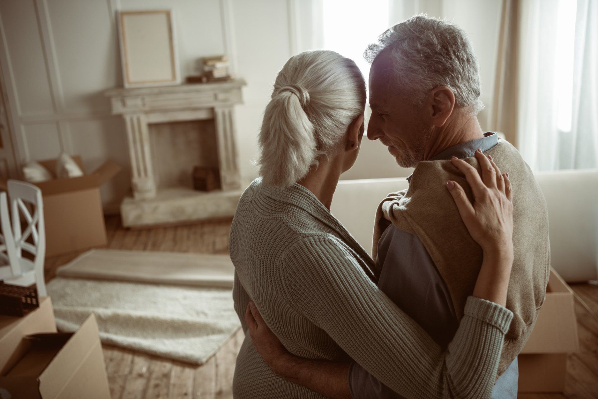 An elderly couple is hugging each other in a living room.