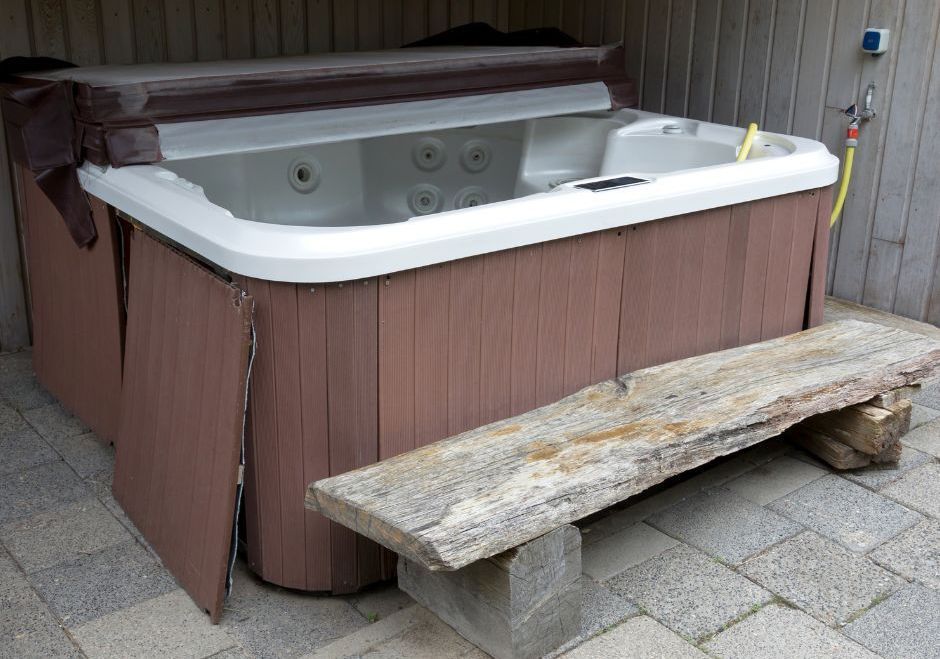 Hot tub with open cover, wooden exterior, bench in front, by a wall.