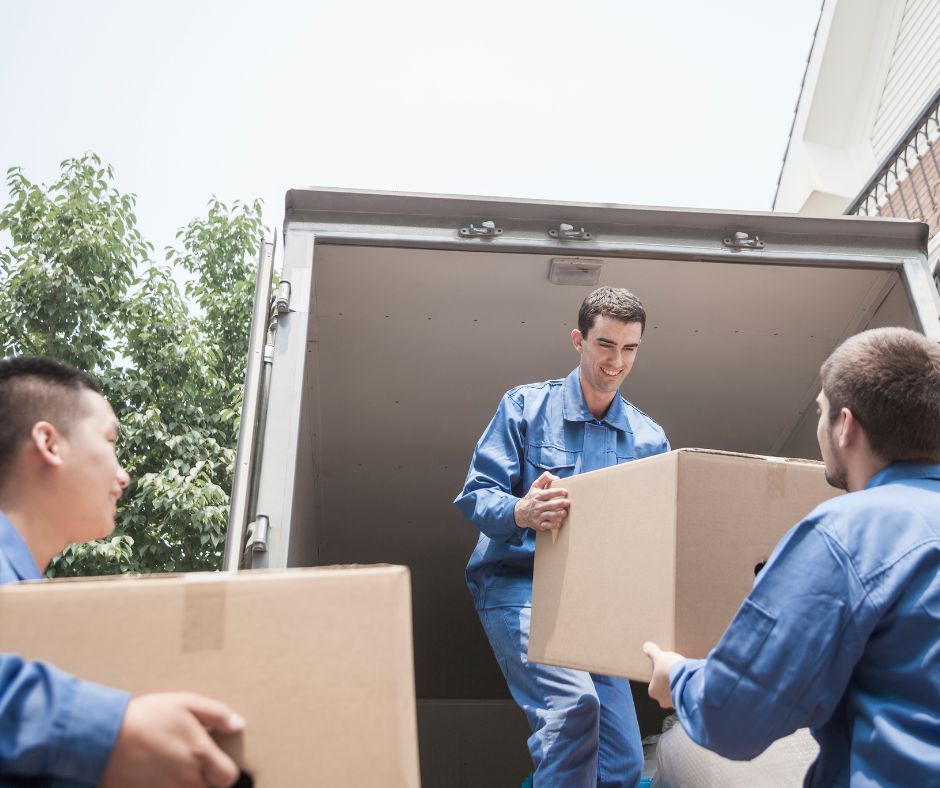 Movers in blue uniforms loading cardboard boxes into a truck, outdoors.