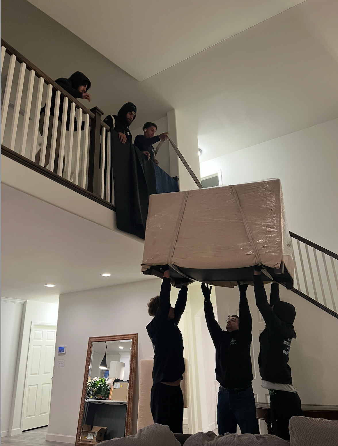Three people carefully moving a piano, wrapped in plastic, in a room with hardwood floors.