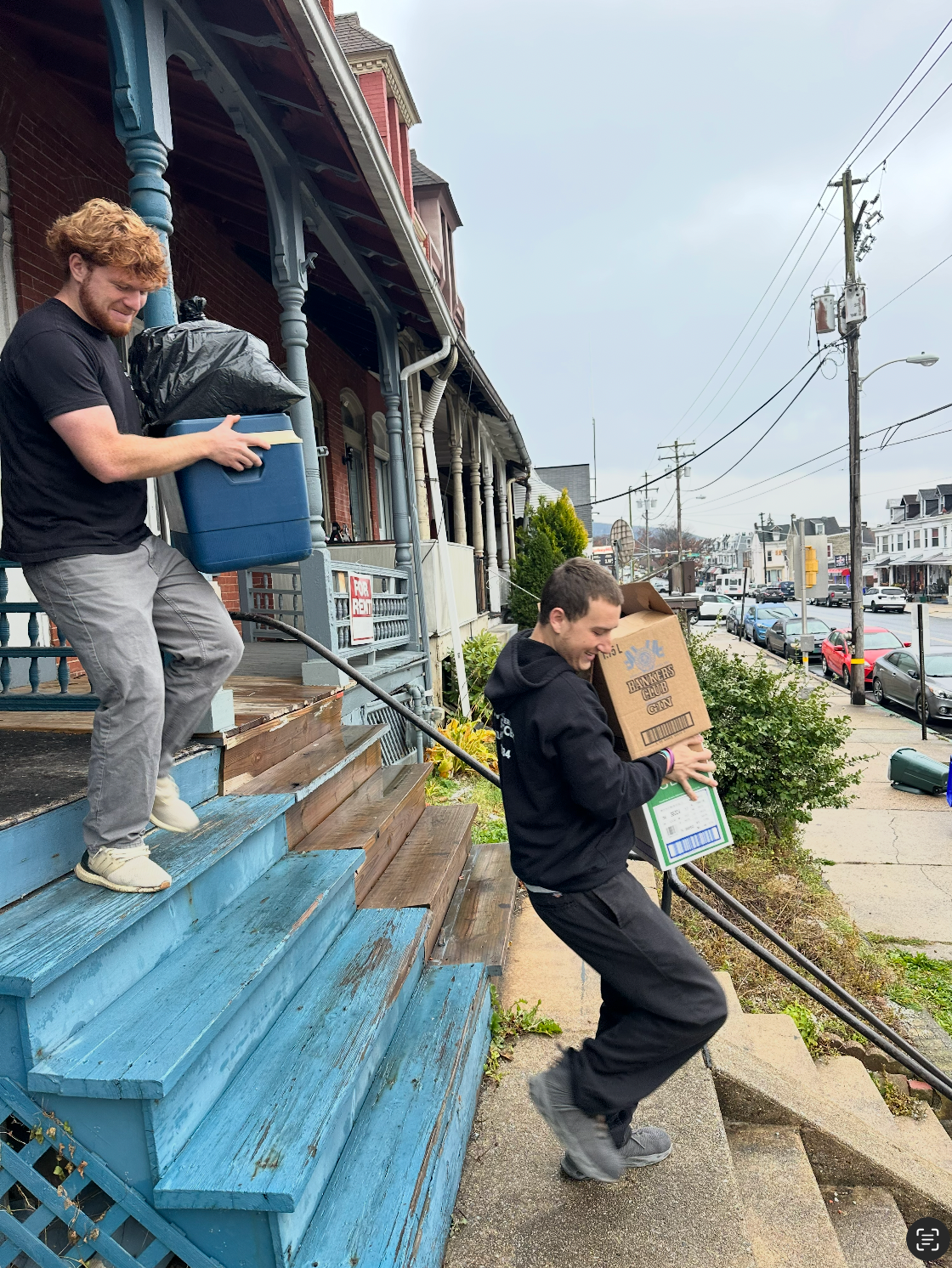 Two people carrying boxes and trash down weathered blue porch steps.
