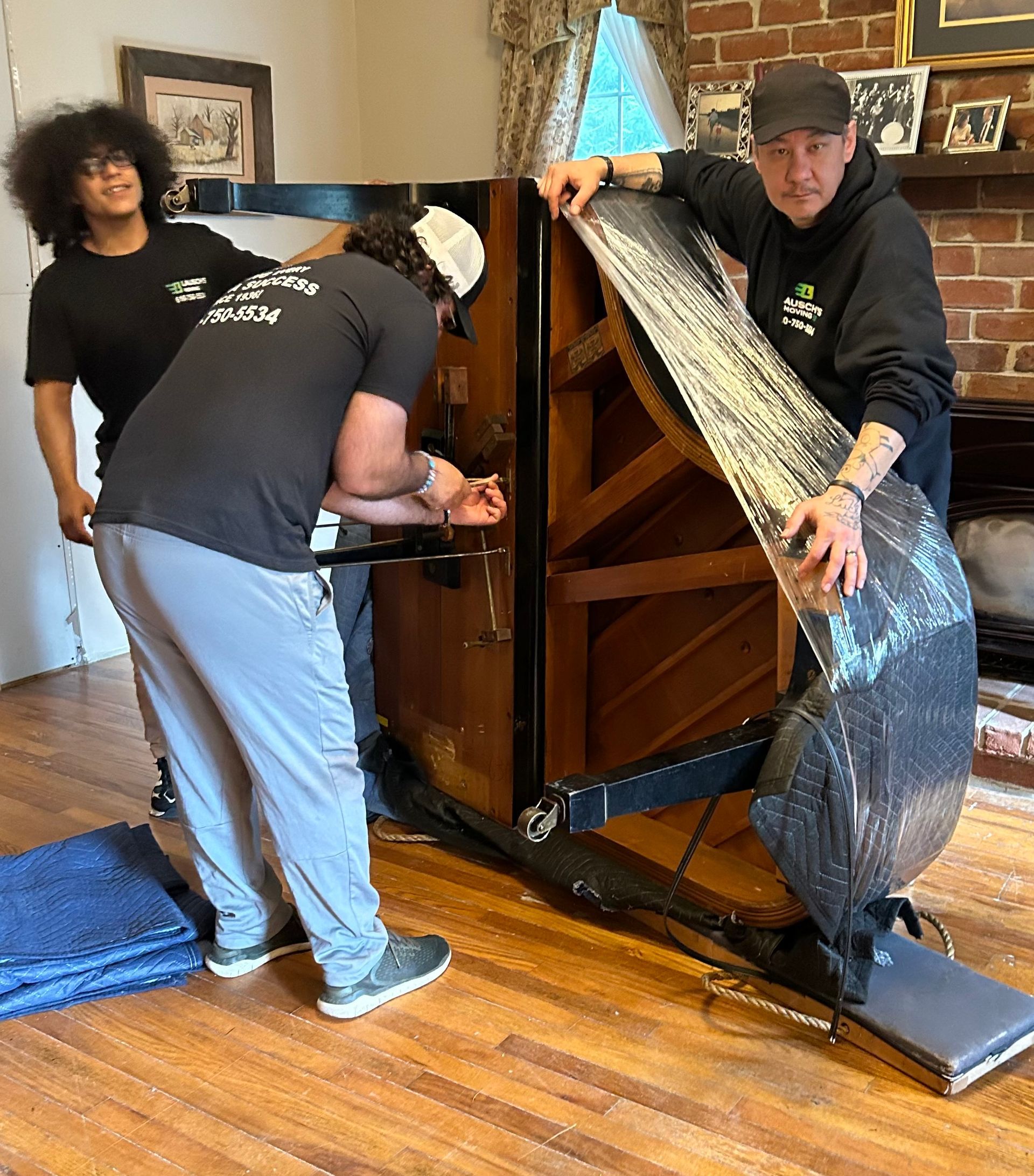 Three people carefully moving a piano, wrapped in plastic, in a room with hardwood floors.