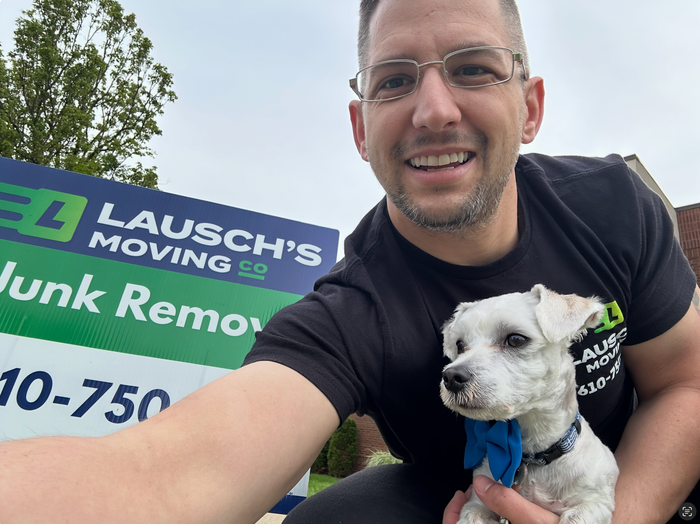 Man in black shirt smiles, holding small white dog with blue bow. Sign reads