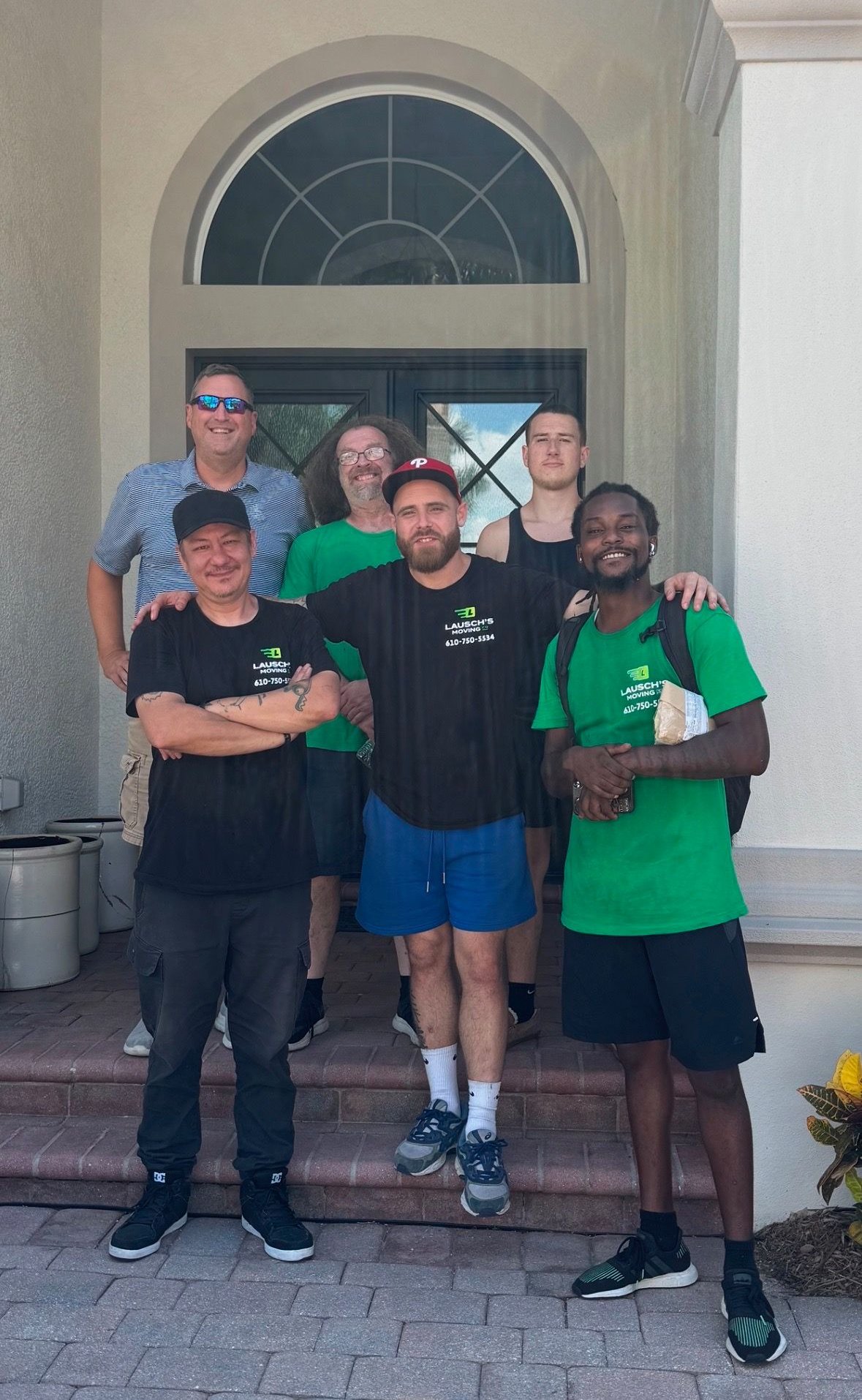 Group of people standing on steps in front of a building with an arched doorway. They wear green and black shirts, smiling.
