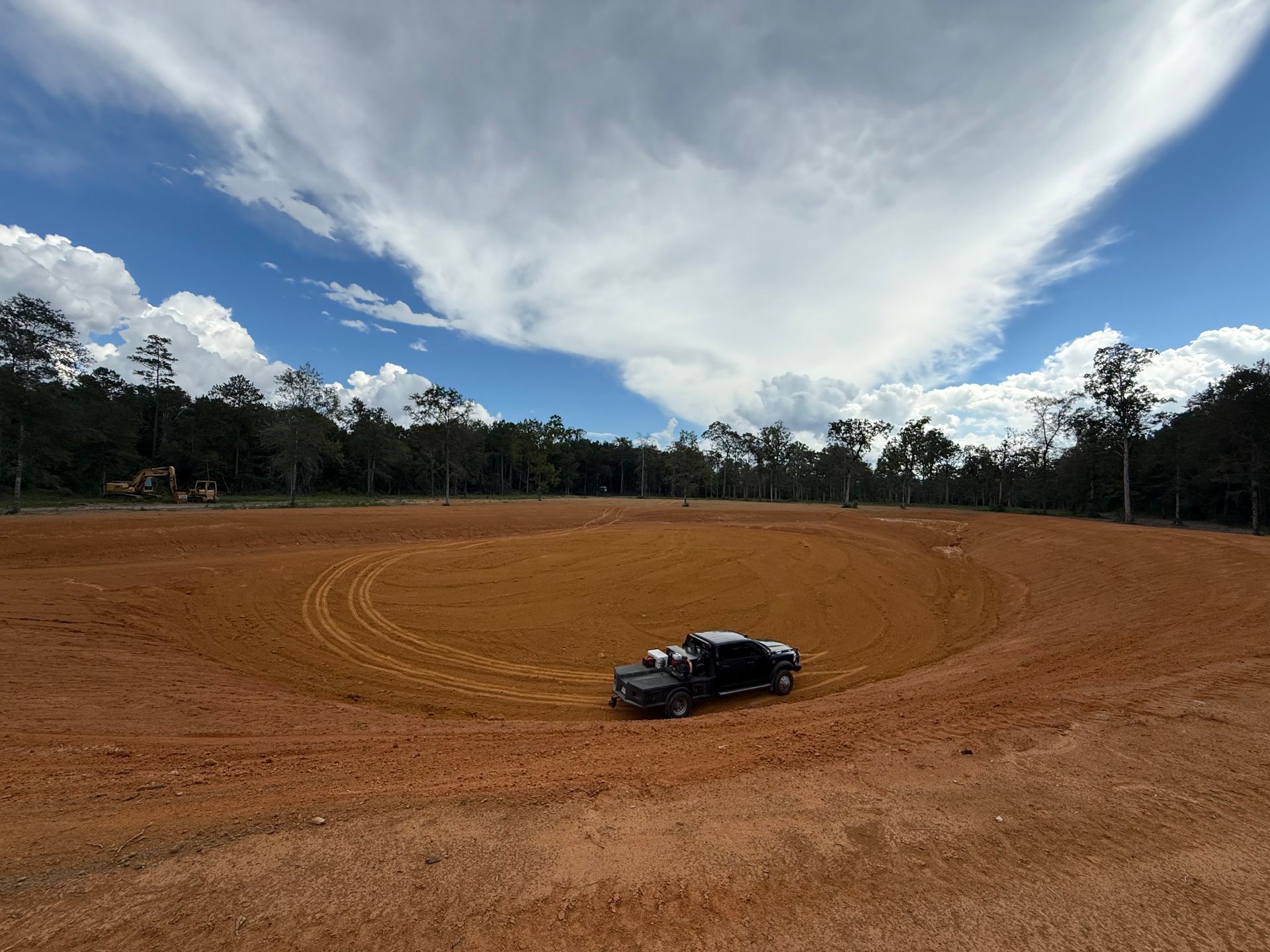 A dark vehicle in a circular dirt track under a cloudy sky. Trees line the background.