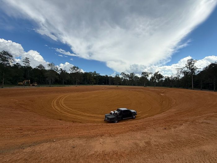 A dark vehicle in a circular dirt track under a cloudy sky. Trees line the background.