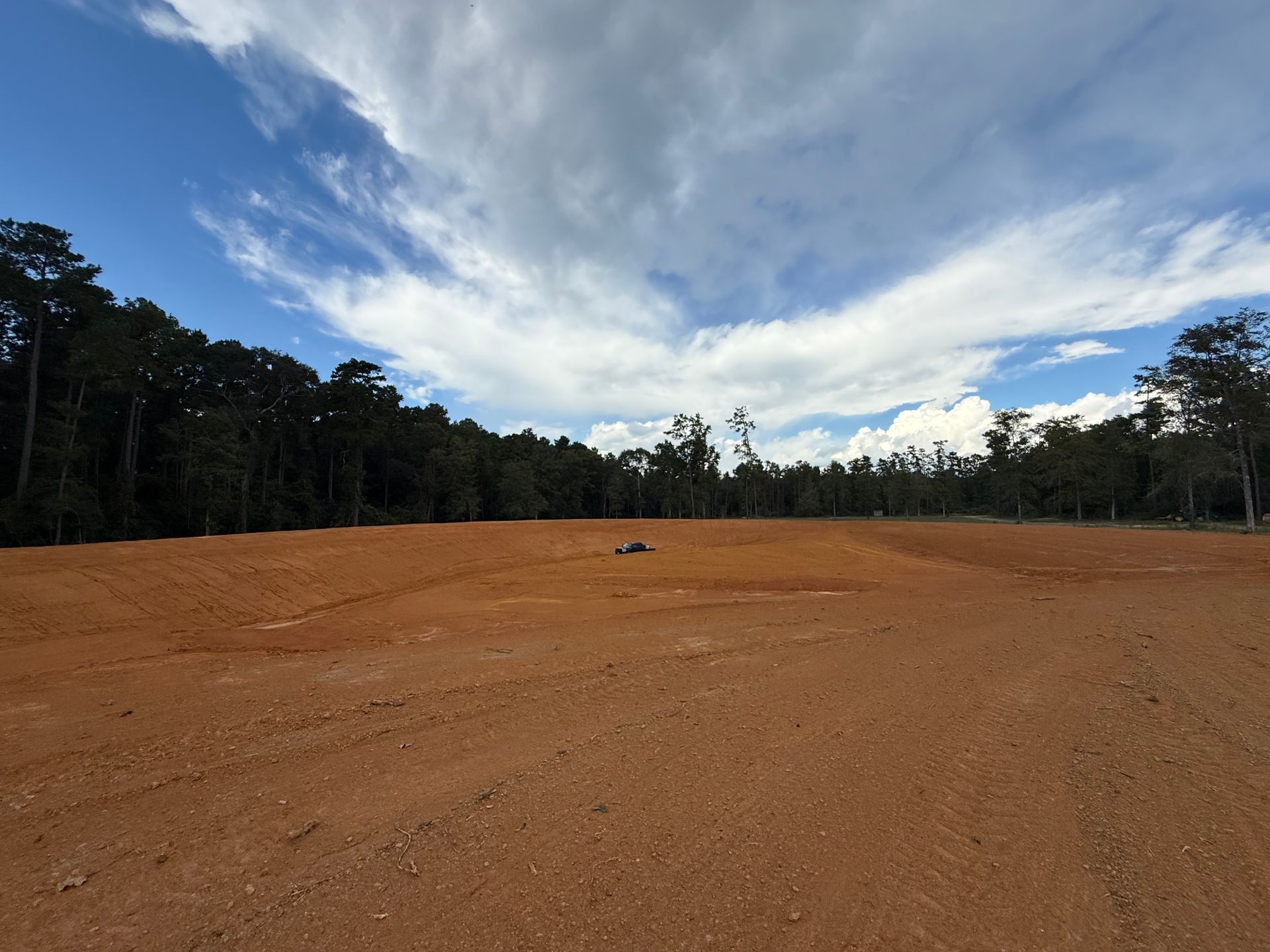 Cleared red dirt field surrounded by trees under a partly cloudy blue sky.