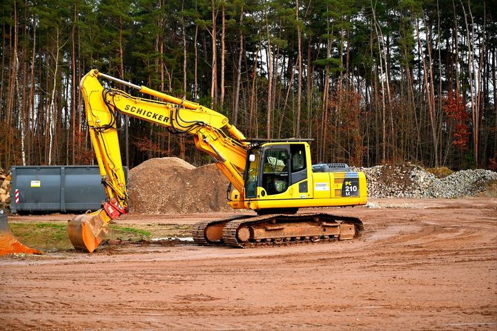 Yellow excavator on a dirt ground in a construction site, near a forest and a pile of dirt.