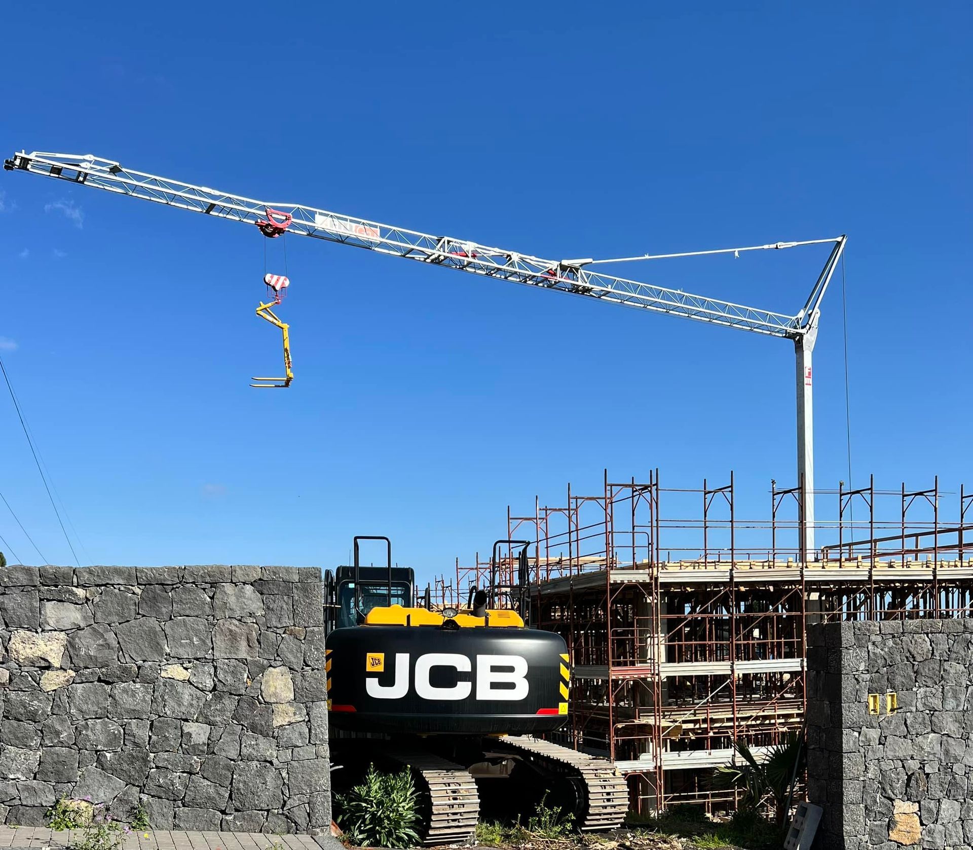 Un cantiere con un escavatore JCB, una gru e un edificio incompiuto sullo sfondo di un cielo azzurro.