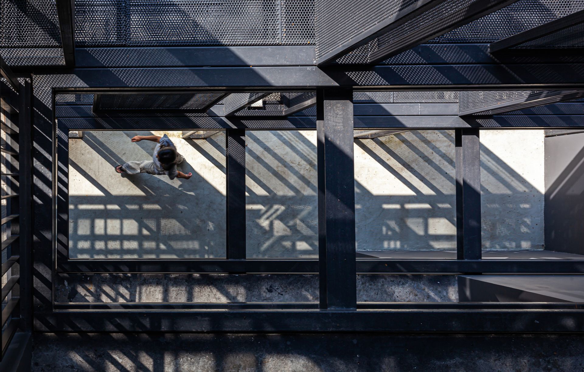 Draufsicht auf eine Person, die auf einer sonnigen Terrasse geht, eingerahmt von einer dunklen Metallstruktur, die Schatten wirft.