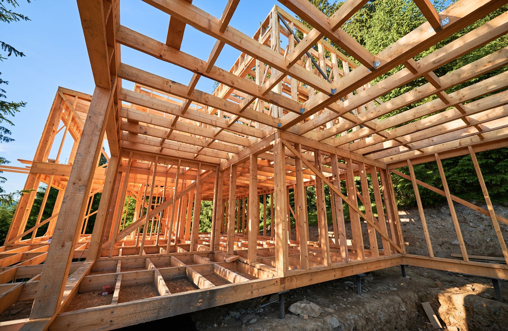 Holzrahmen eines im Bau befindlichen Hauses vor blauem Himmel mit viel Grün im Hintergrund.