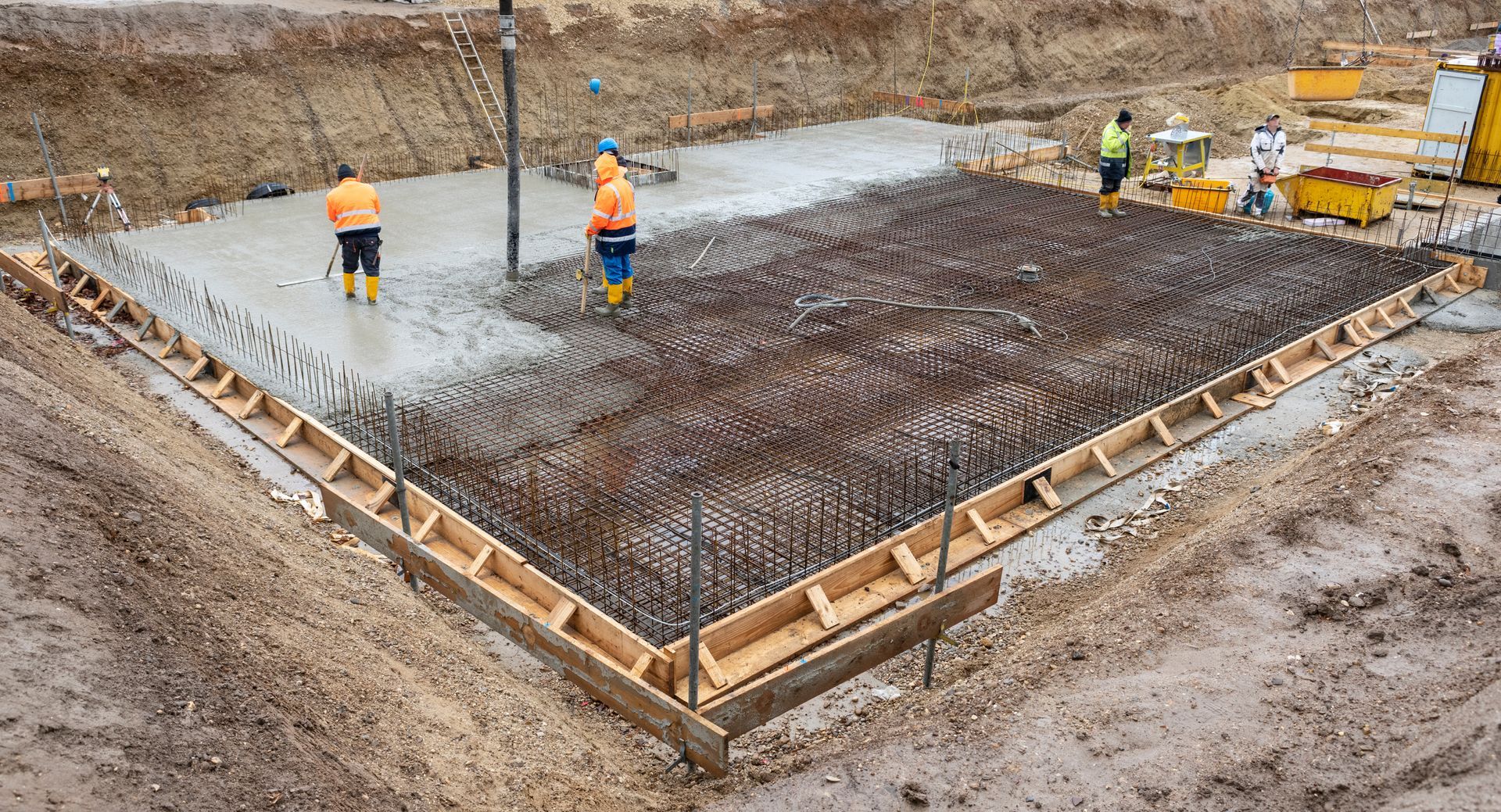 Baustelle: Arbeiter gießen Beton über Bewehrungsstäbe in Holzschalungen; umgeben von Erde.