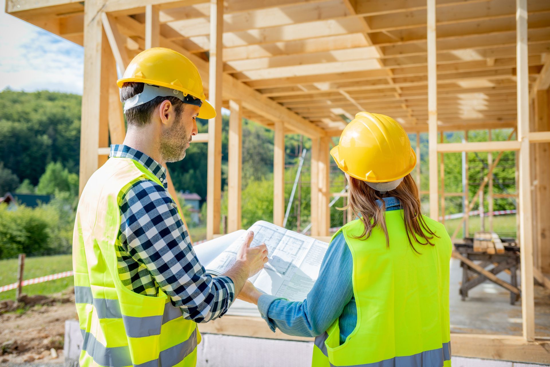 Zwei Bauarbeiter prüfen auf einer Baustelle mit Holzrahmenbauwerk die Baupläne. Sie tragen Sicherheitswesten und Schutzhelme.