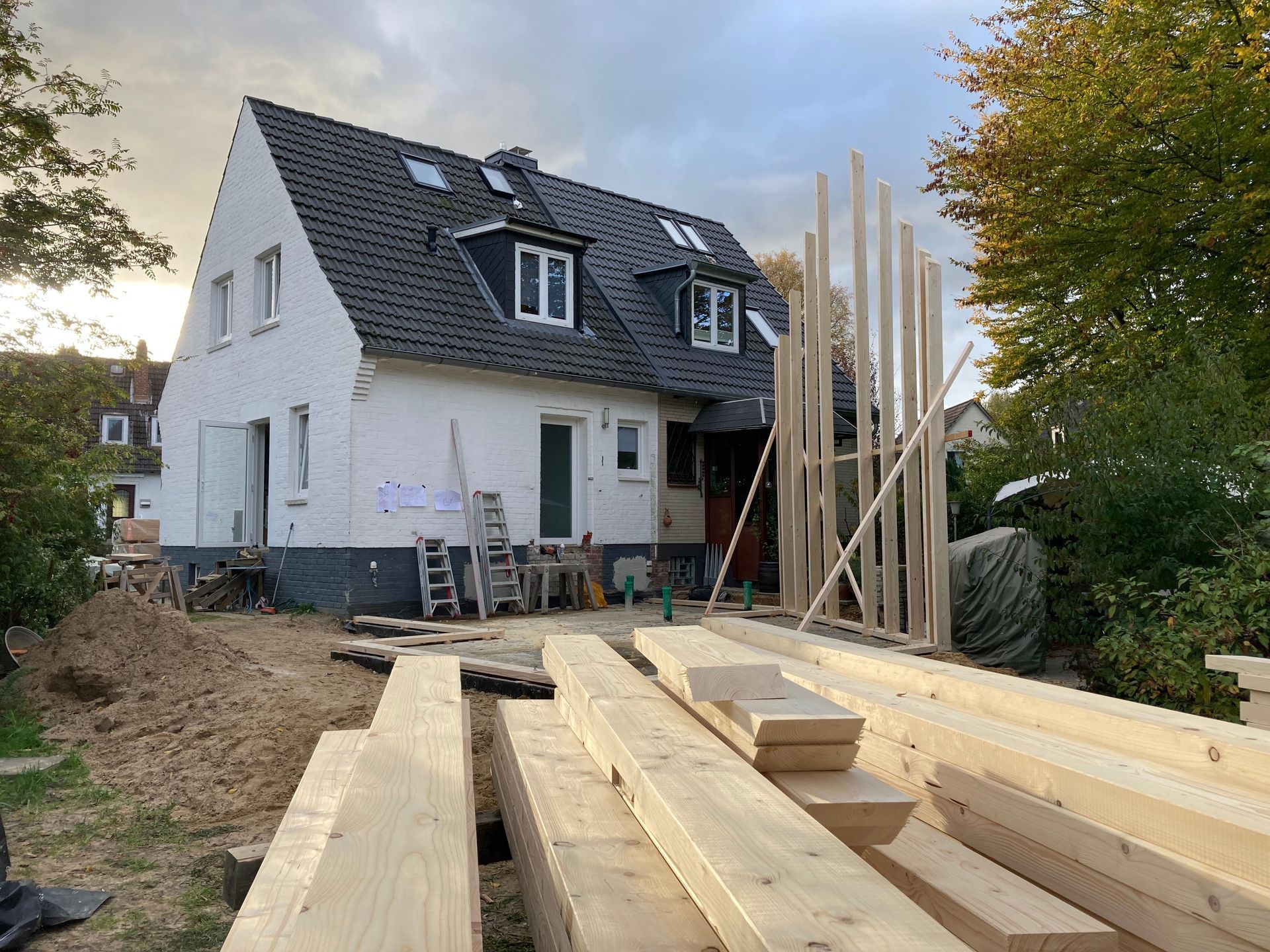 Haus im Bau; davor Bauholz und Holzbalken. Weiße Außenseite, schwarzes Dach und hohe Holzbalken.