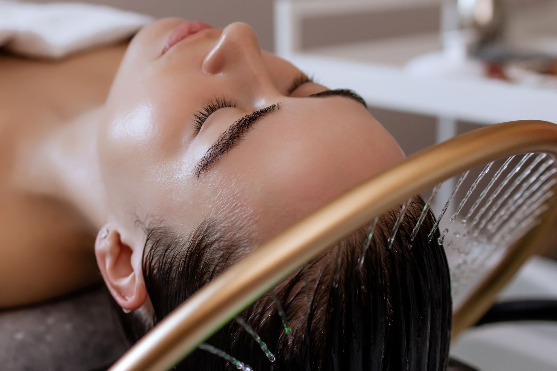 Person reclining with wet hair being rinsed at a salon sink, water pouring over the scalp