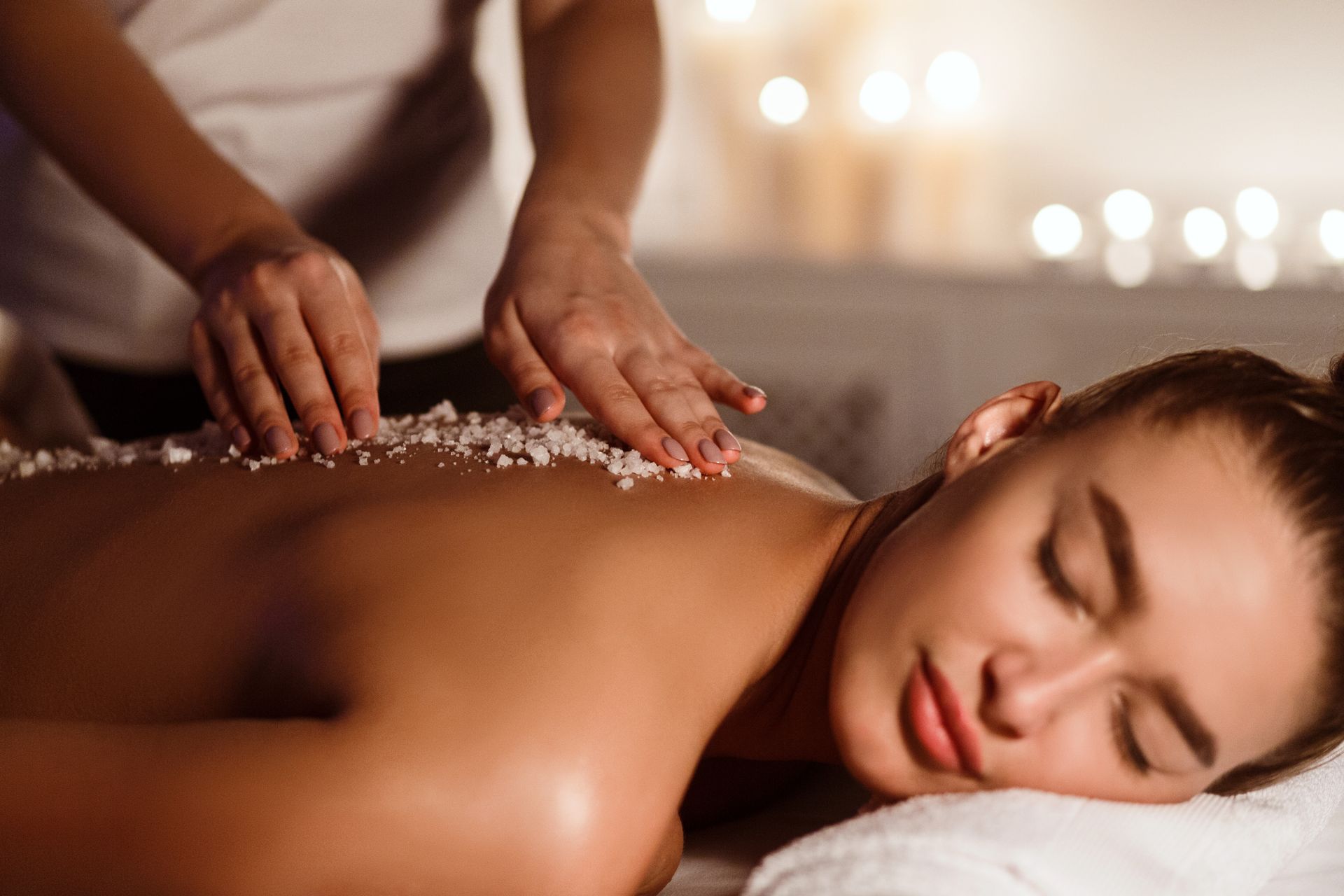 Spa massage with hands applying salt scrub to a woman’s back as she rests on a towel