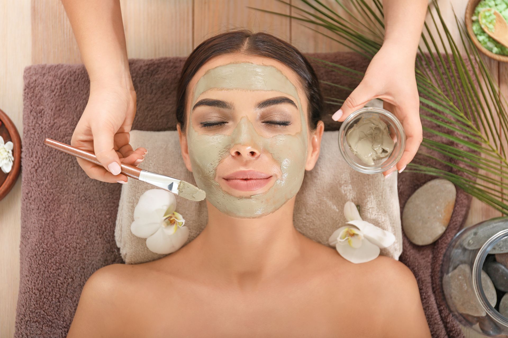Woman receiving a green facial mask during a spa treatment with brushes, bowls, and flowers nearby