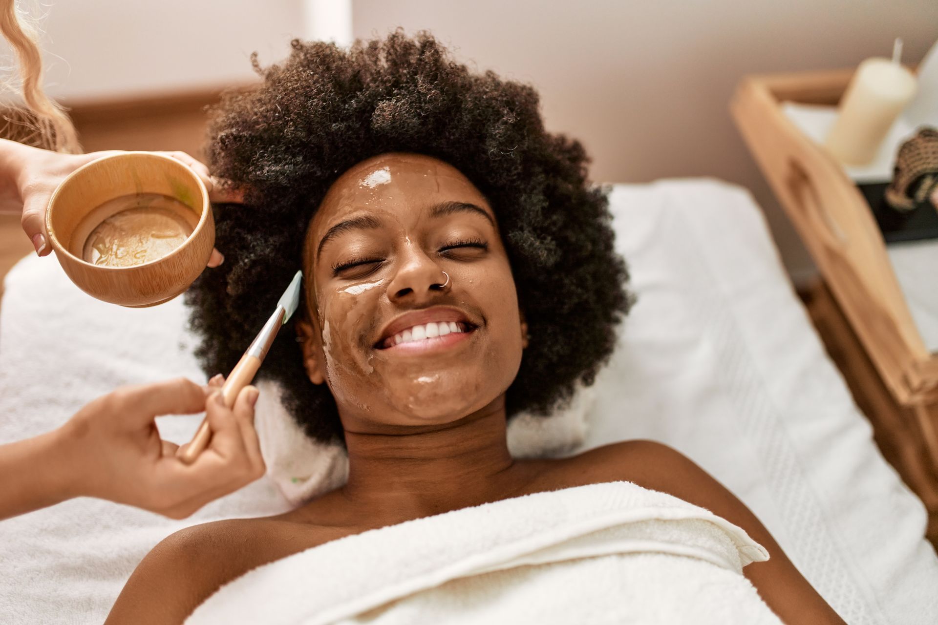 Woman smiling during a facial treatment, with cream applied and a brush and bowl nearby.