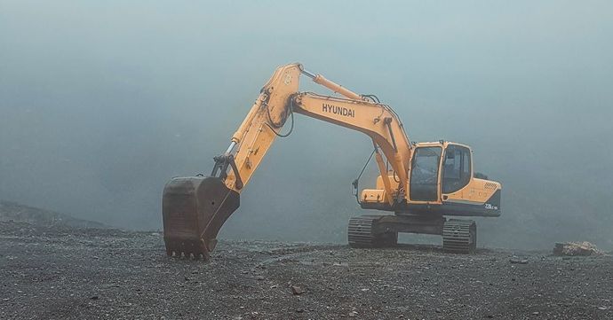 A yellow excavator is working on a rocky hillside in the fog.