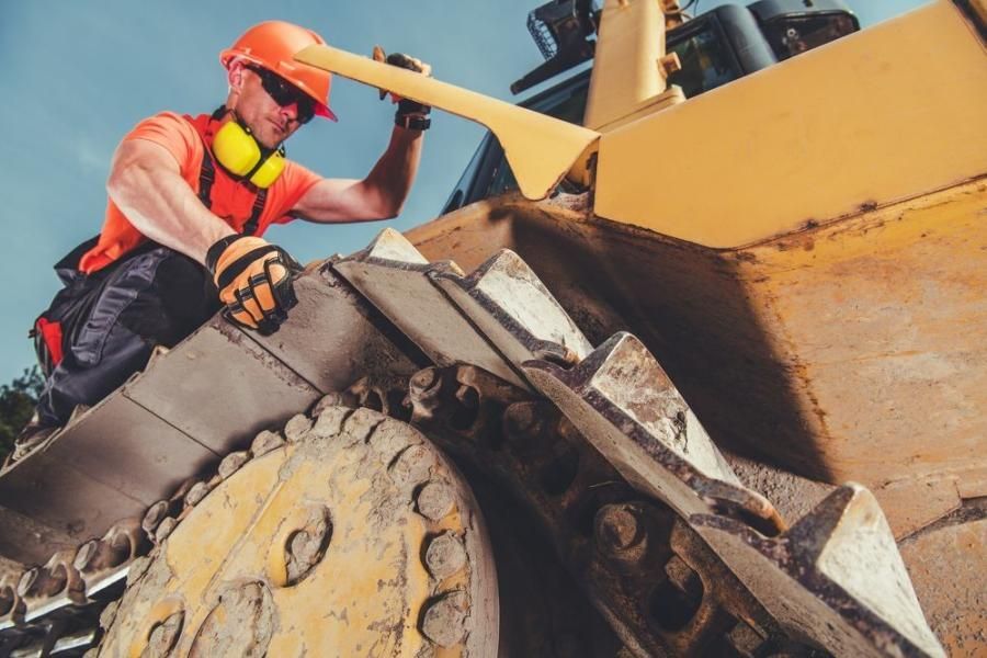 Construction worker examining a bulldozer track; wearing orange safety gear; outdoors.