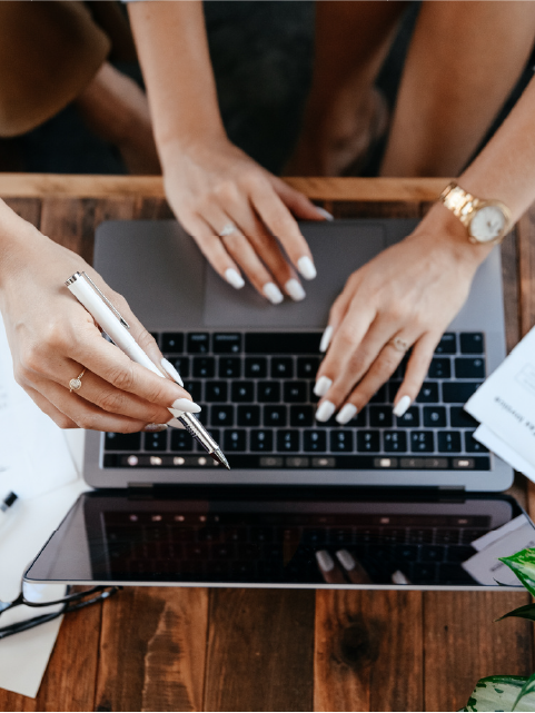 Hands on laptop, one pointing pen, wooden desk.