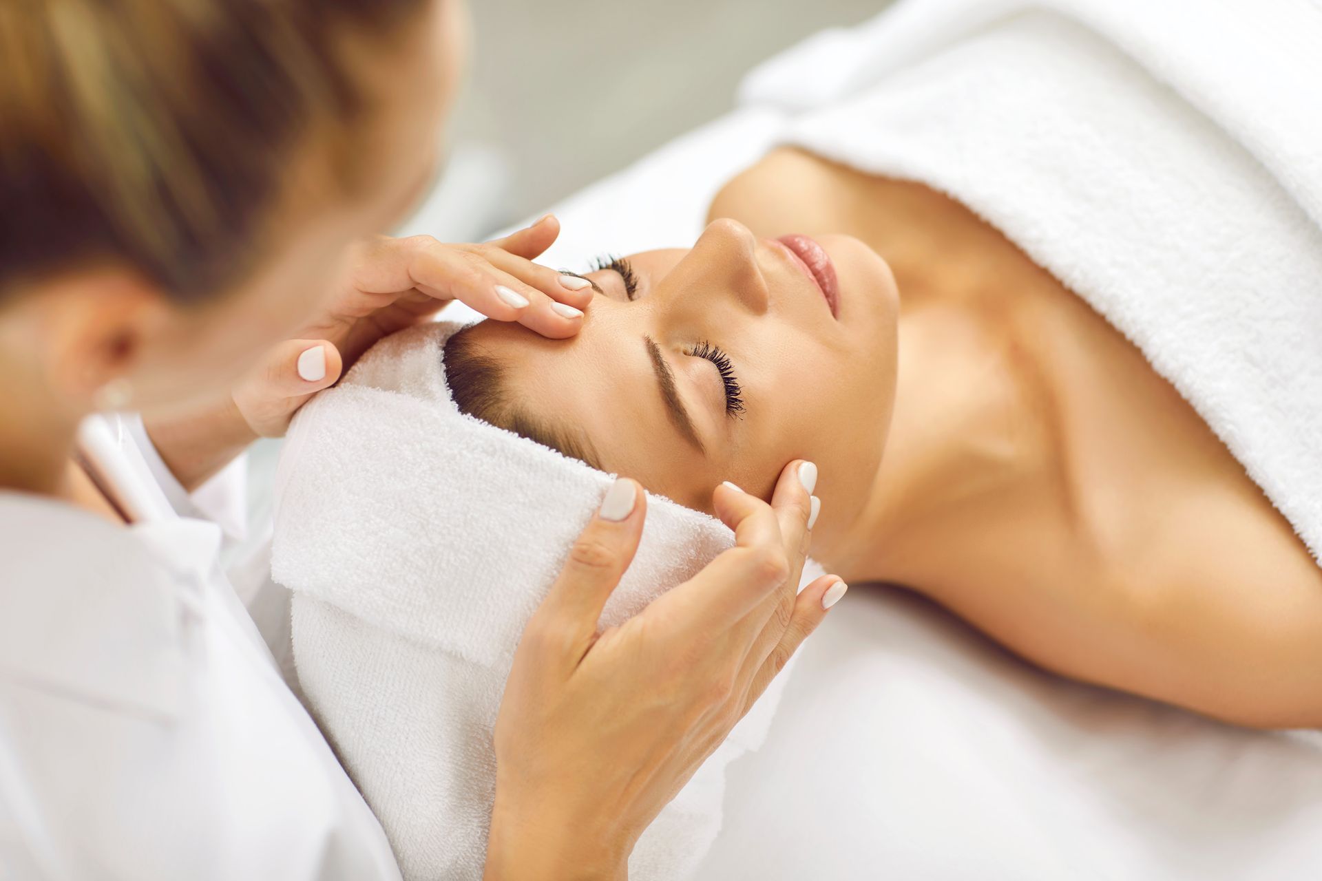 A spa facial treatment with a woman receiving a forehead massage and wearing a towel headwrap