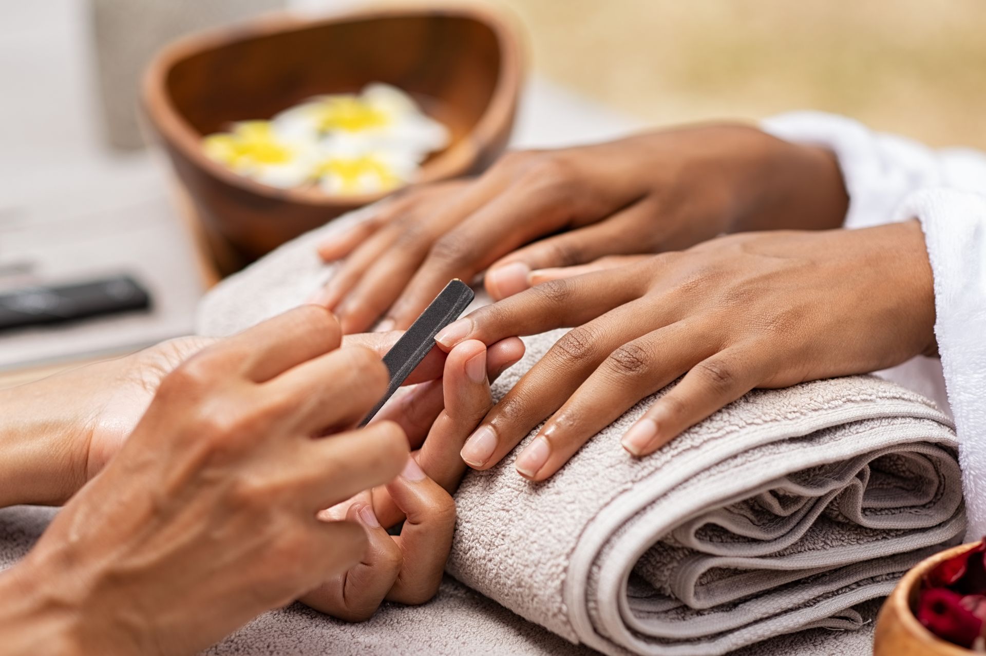 Manicurist applying nail polish to a client’s hand beside rolled towels and a bowl of flowers.