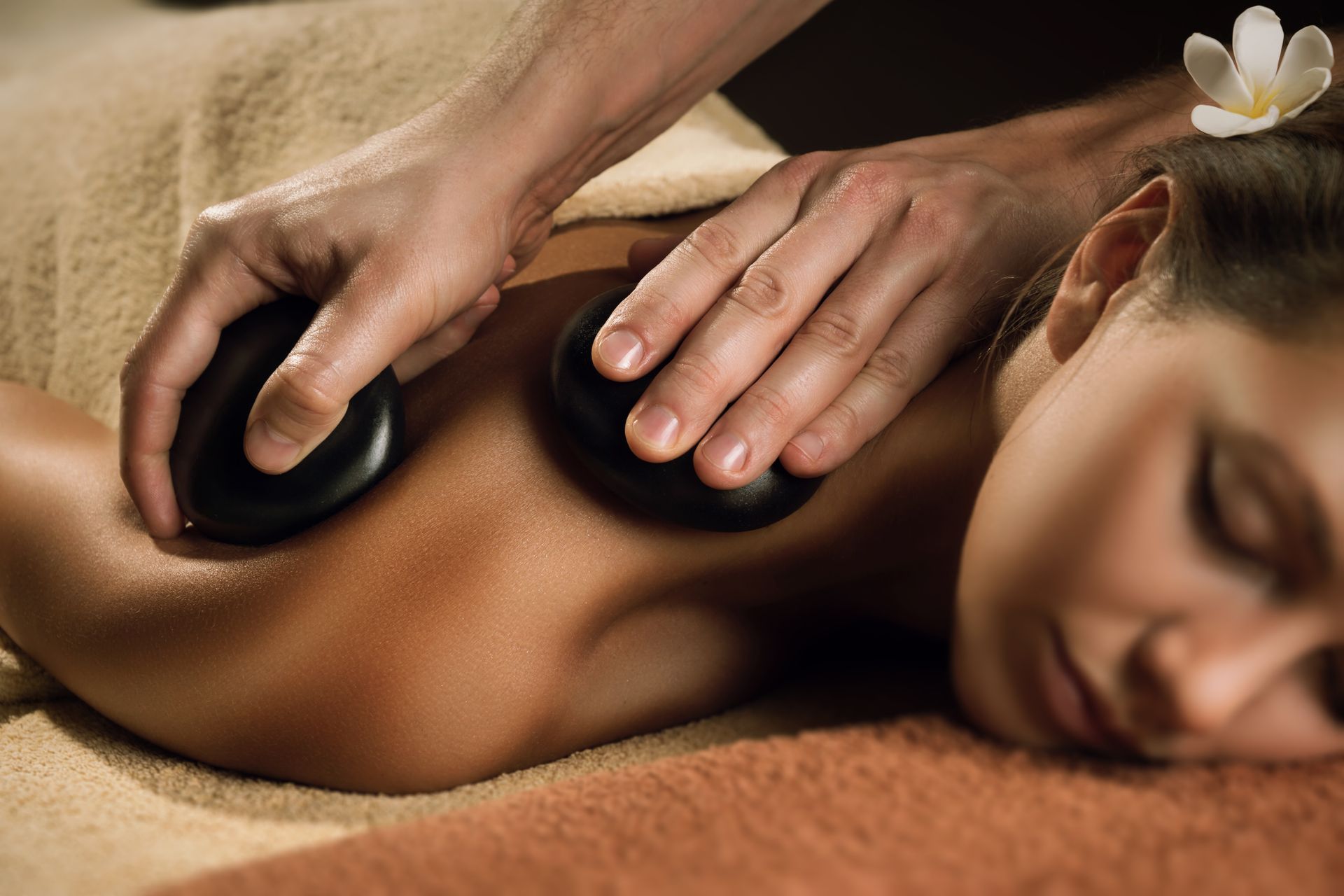 Person receiving a hot stone massage on a spa table with closed eyes and white flowers nearby