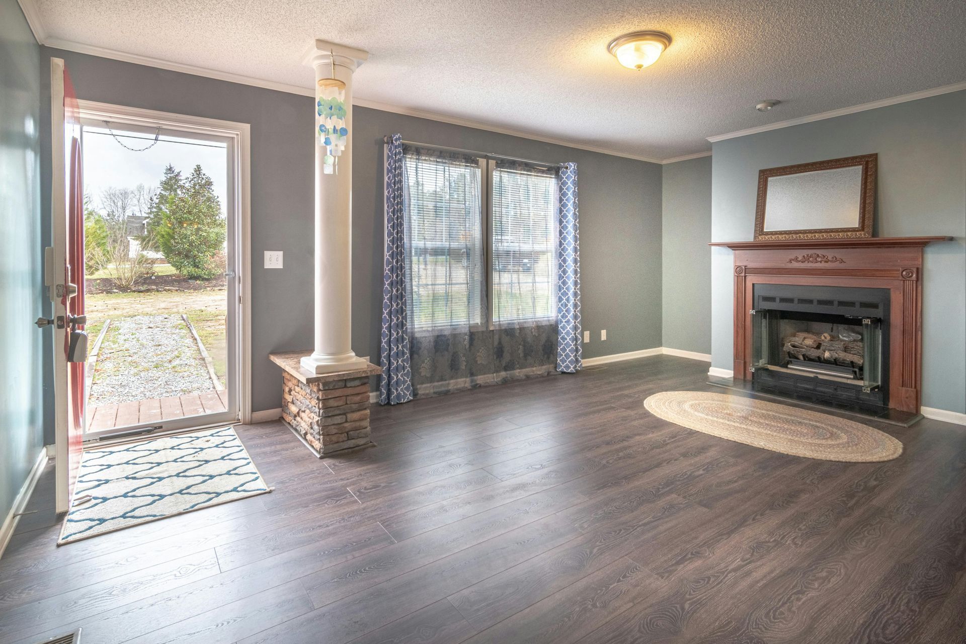 An open living room with a fireplace, dark wood floors, a decorative column, and a door leading to an outdoor path.