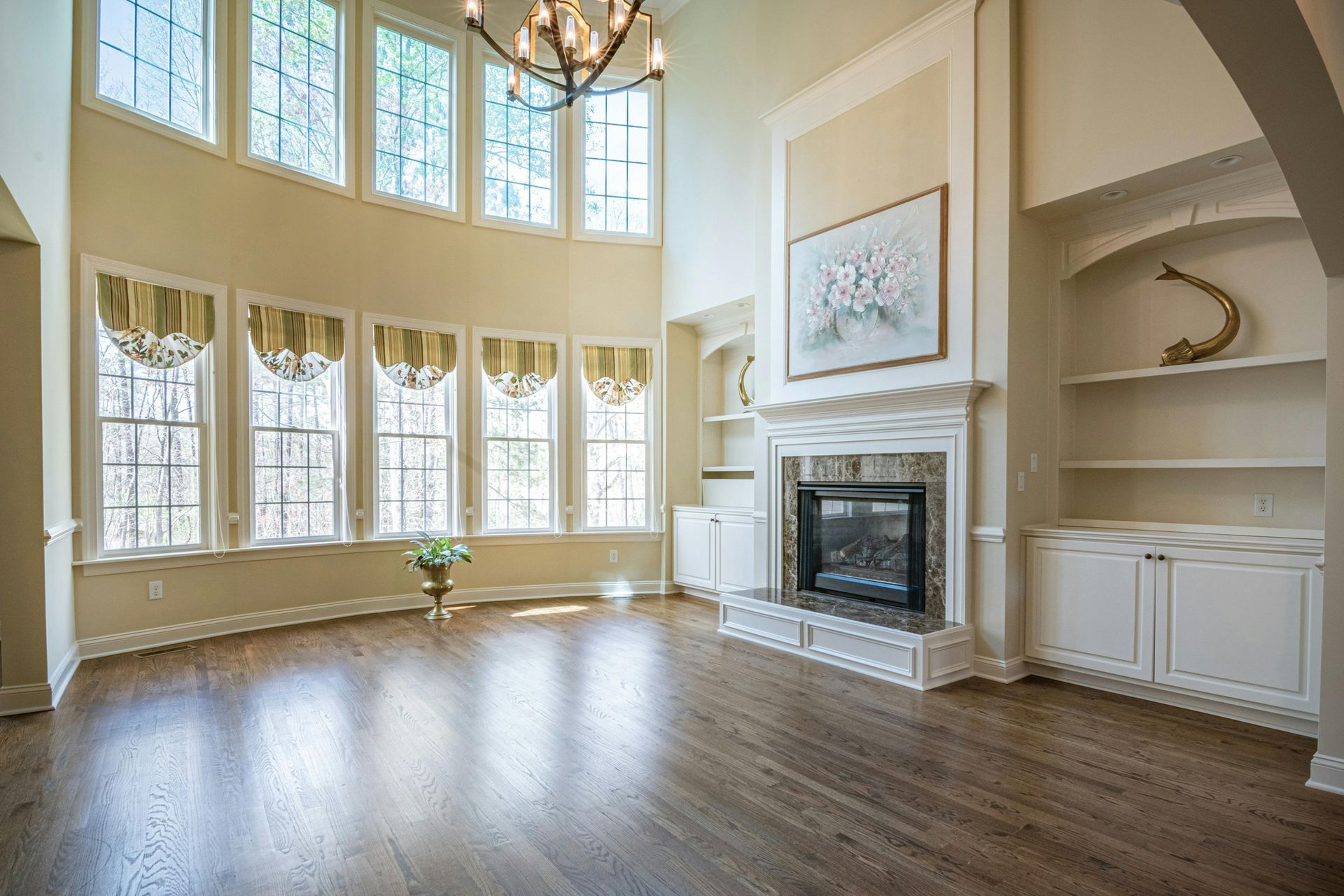 A spacious living room with hardwood floors, a stone fireplace, built-in shelving, and large windows with valances.