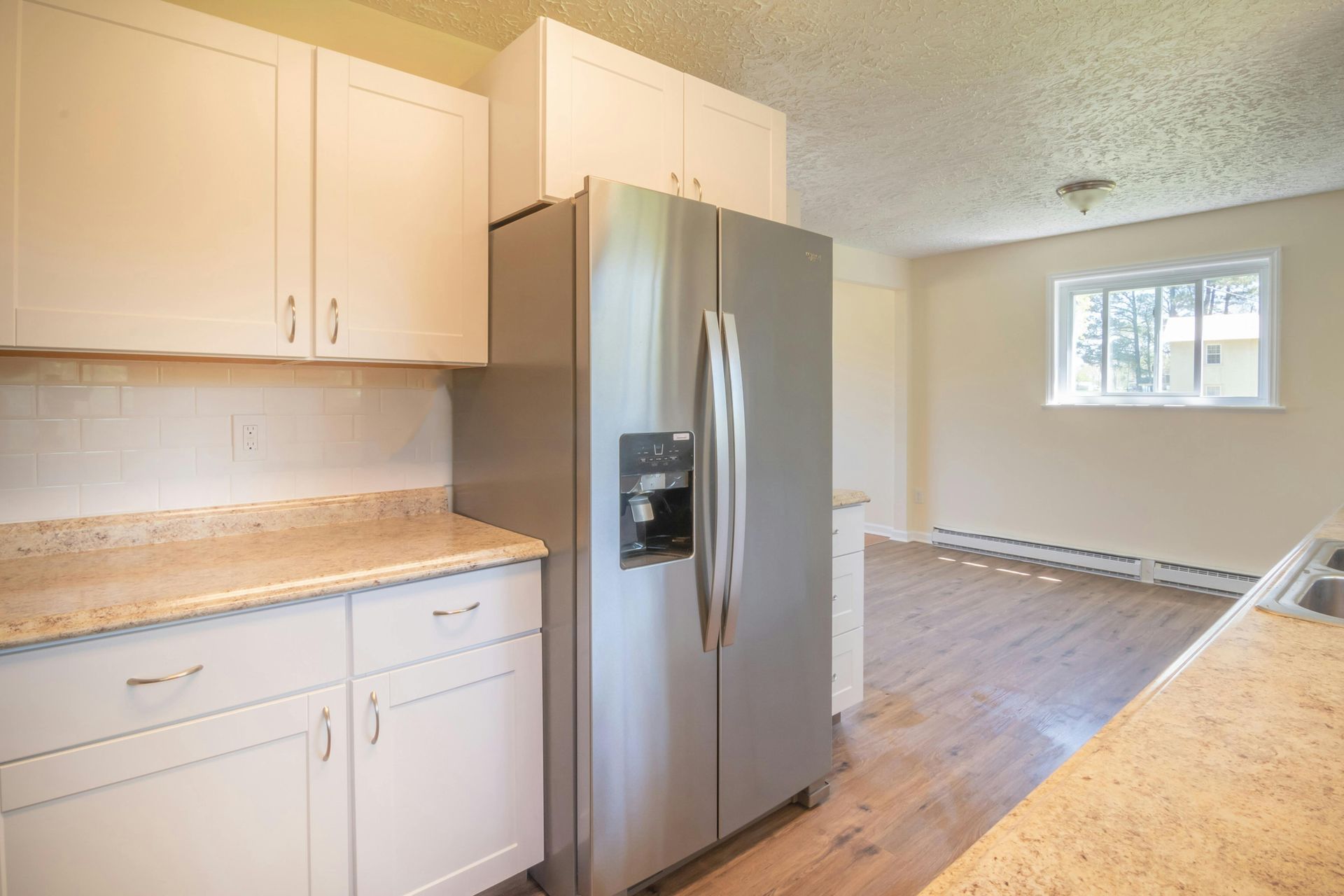 A bright kitchen with white cabinets, speckled countertops, stainless steel refrigerator, and view of an adjacent room.
