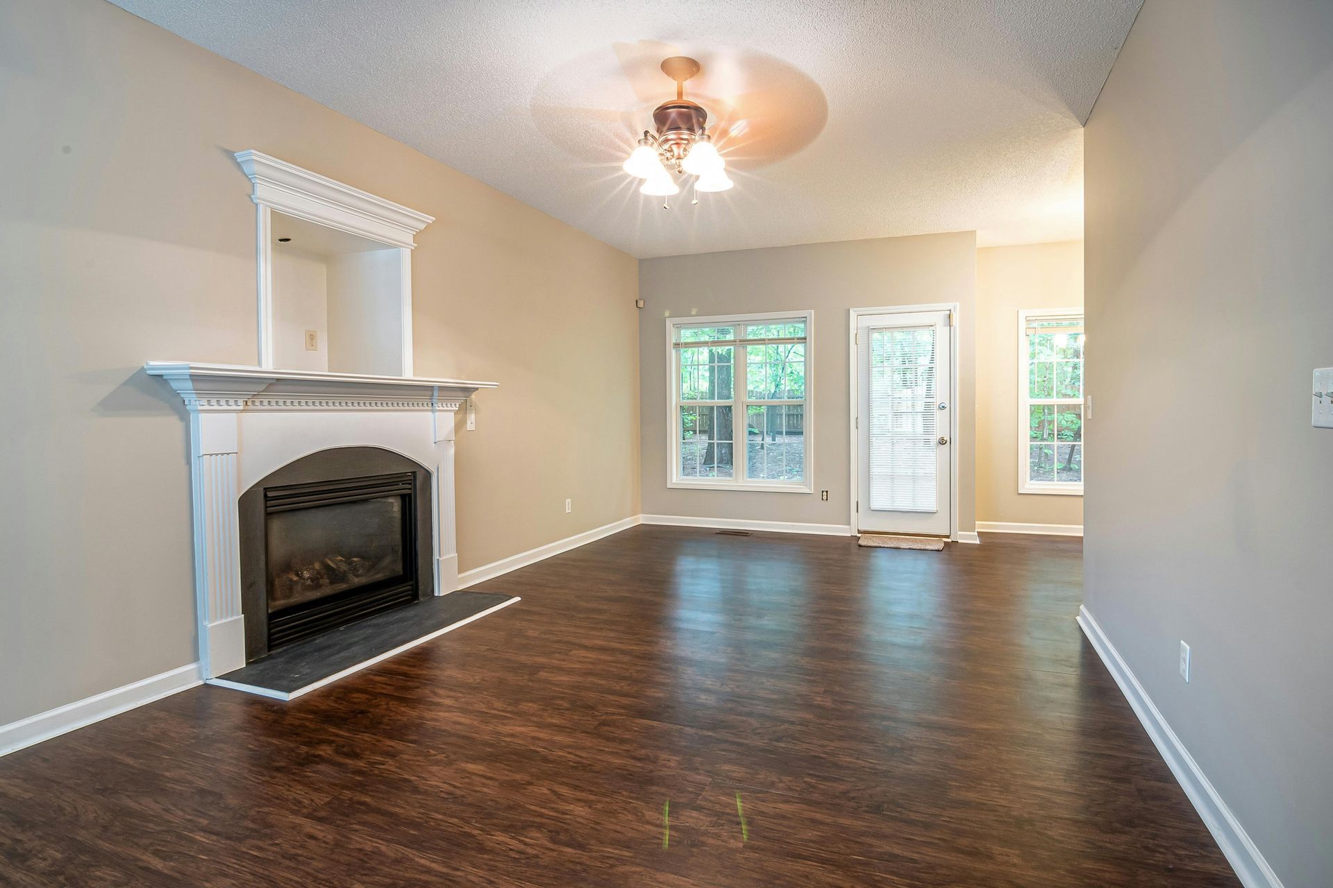 A spacious living room with dark wood floors, beige walls, a white fireplace, ceiling fan, and a door leading outside.