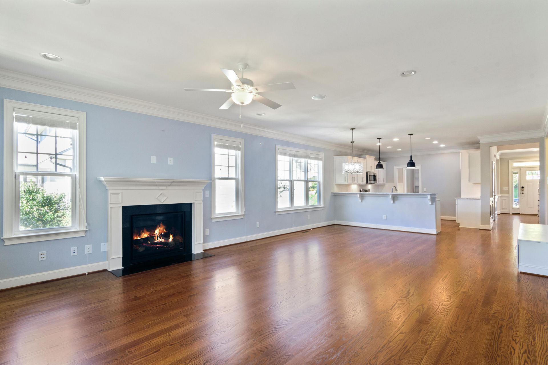 An open-concept living area with light blue walls, hardwood floors, a gas fireplace, and a kitchen visible in the back.