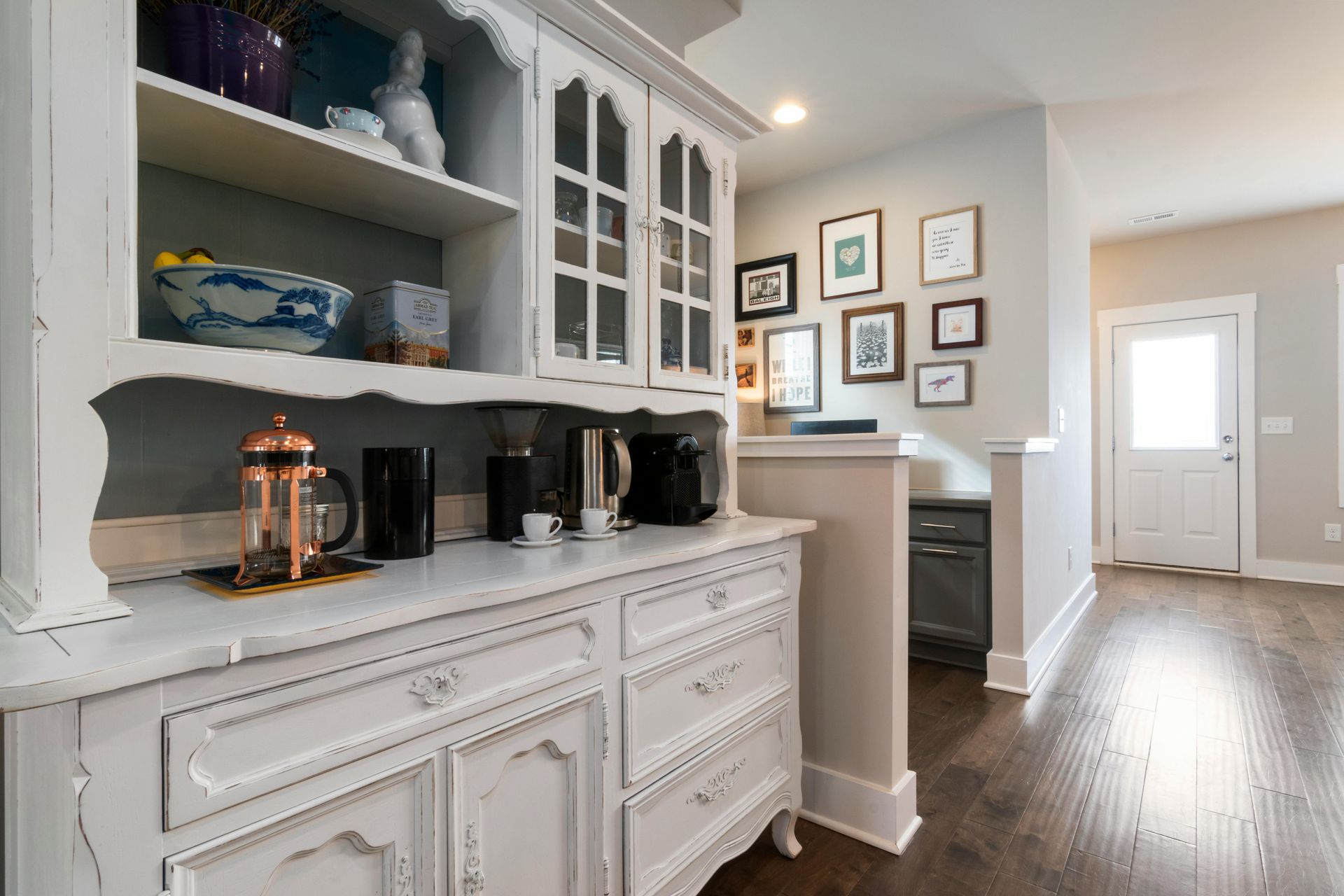 A white antique hutch filled with kitchen items sits in a room with a gallery wall, hardwood floors, and a white doorway.
