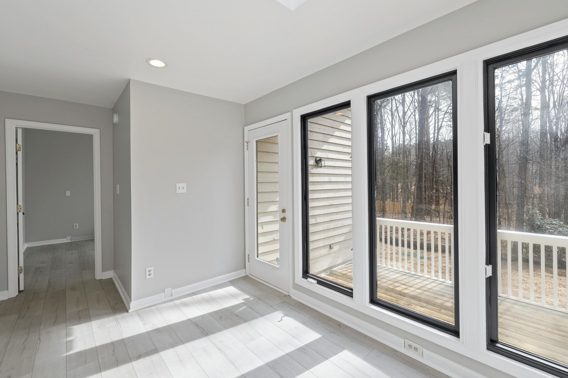 A bright, modern sunroom with light gray walls, wood-look flooring, a glass-paneled door, and large windows facing a deck.