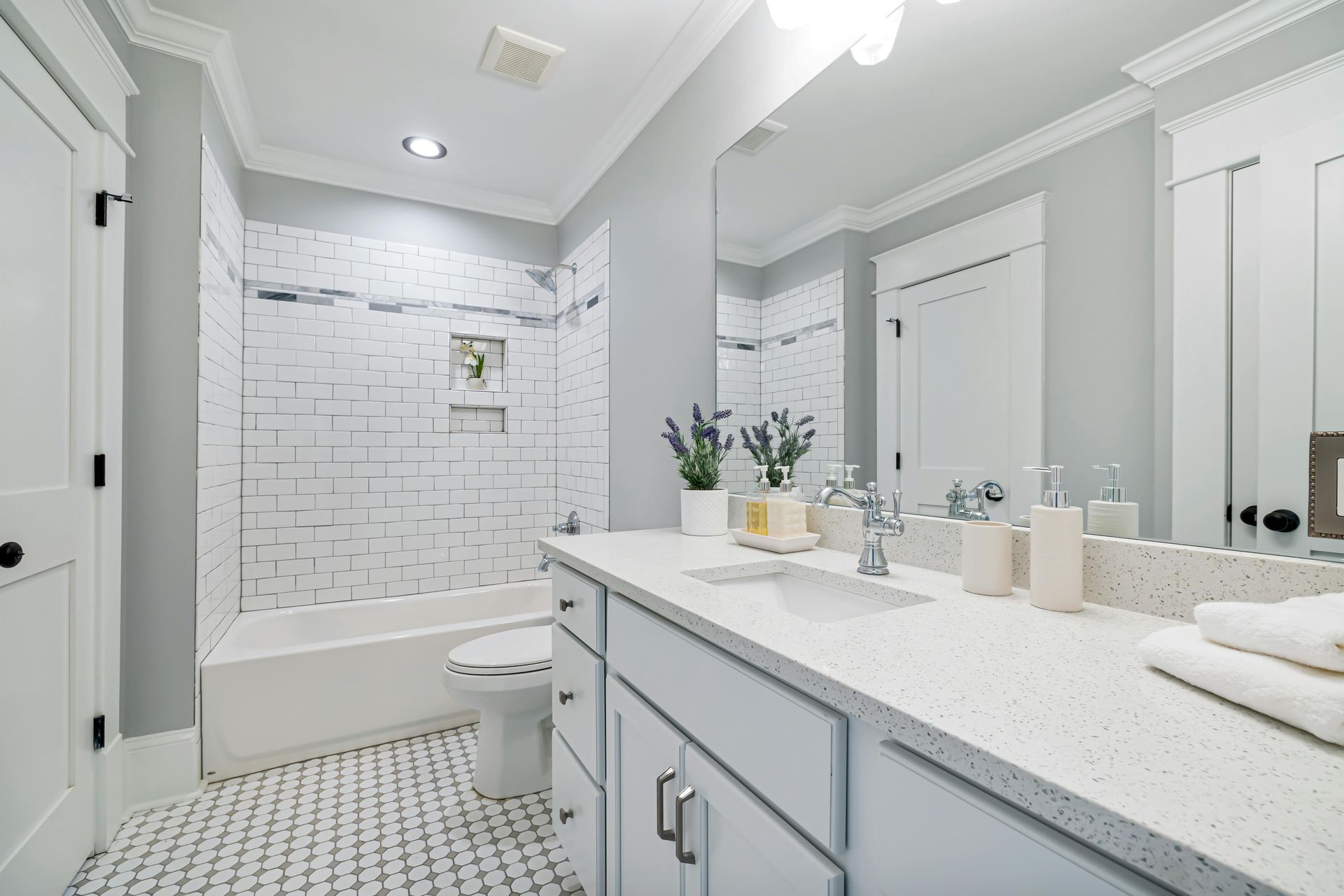 A bright, modern bathroom with a white tiled tub, gray walls, light countertops, and a large vanity mirror.