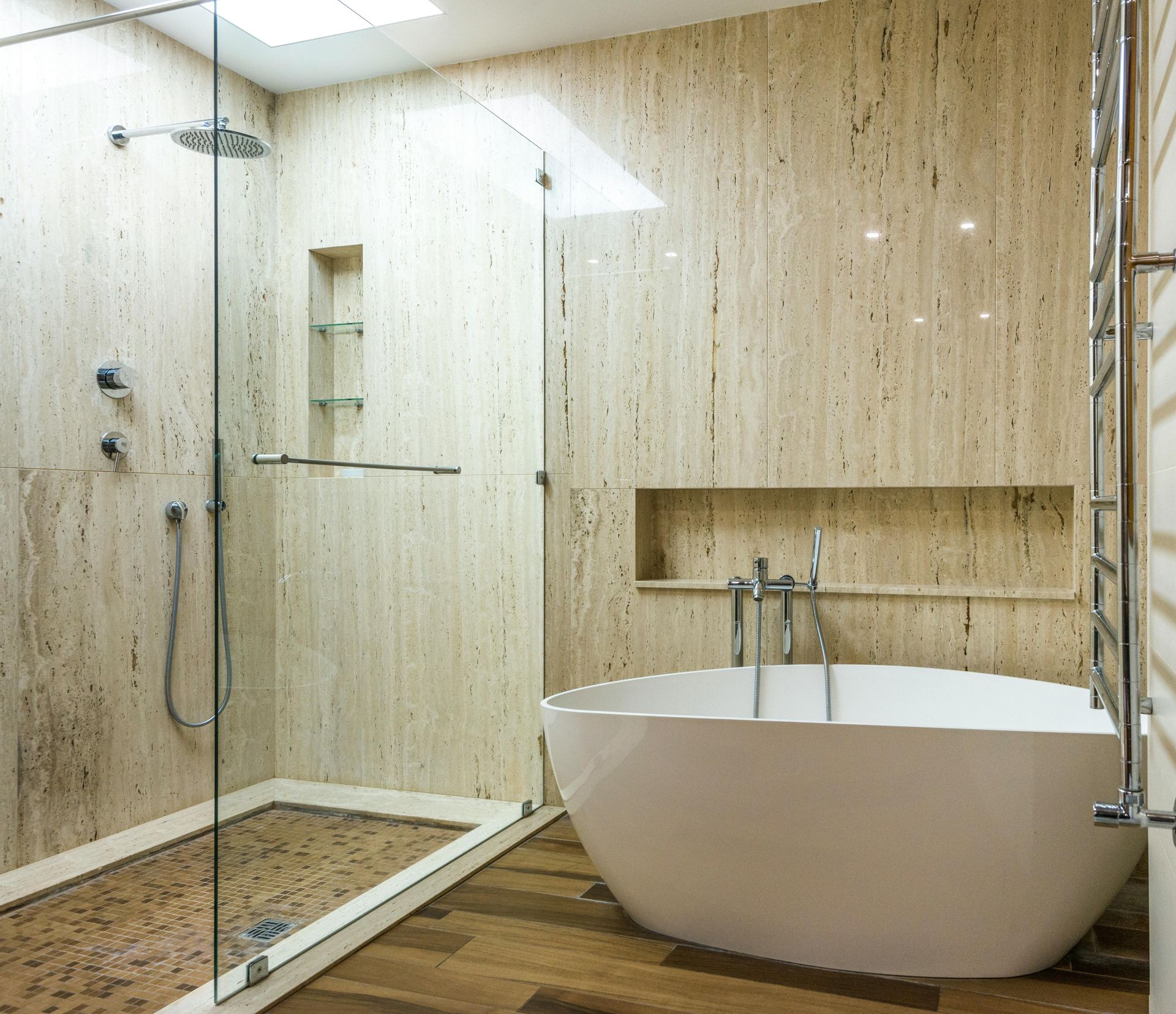 A modern bathroom with a glass-enclosed shower, a white freestanding tub, and beige stone tile walls.